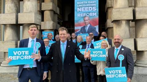 A group of people standing outside Grimsby Town Hall wearing office attire, holding blue "VOTE REFORM" signs. There is a large banner behind them featuring an image of Nigel Farage below the writing "NORTH EAST LINCOLNSHIRE NEEDS REFORM". The party's deputy leader, Richard Tice, is standing in the middle of the group, smiling, with his arms open. 