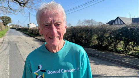 Mike, who is wearing a bright blue T-shirt with the words: Bowel Cancer UK, is smiling at the camera during a summer's day. 