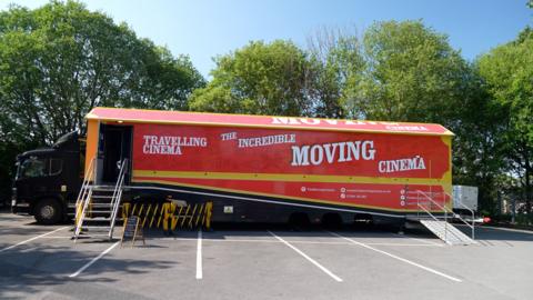 A lorry parked in a car park in front of some trees. The red lorry has the words 'travelling cinema' and 'the incredible moving cinema' on the side. A set of stairs up to a door into the lorry.