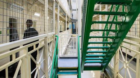 Prison wing showing a staircase and a female prison officer escorting an inmate to his cell