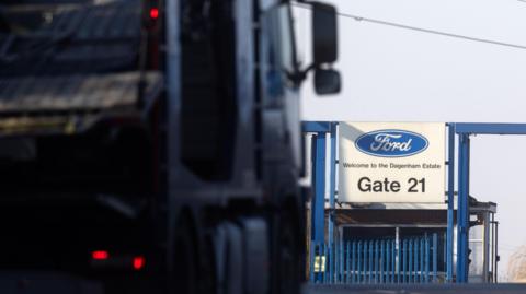 A lorry approaches the bright blue security fence of Ford car plant at Dagenham. There is a big white sign with the blue Ford logo which states "Welcome to the Dagenham Estate - Gate 21".