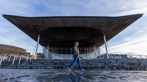The Senedd building is a glass structure with a large wooden roof which overhangs slate steps that cascade down from the building to a walkway below.