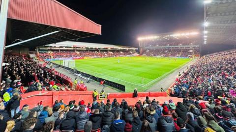 Pittodrie Stadium - Aberdeen's stadium, full of fans, around a green football pitch, under floodlights and dark skies.