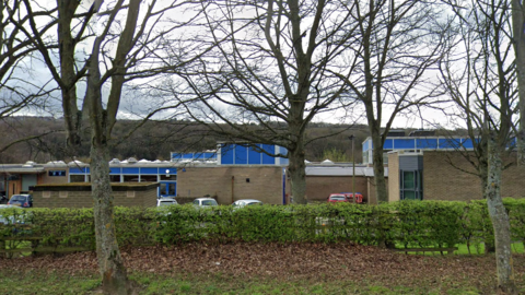 The exterior of Ovingham Middle School which is a brown building with blue details. In front of the building is a car park and grass with leafless trees.