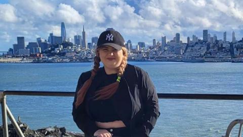 June Bunyan photographed with the city of San Francisco in the background. She's wearing a black basketball cap and a black jacket. The water is in the background.
