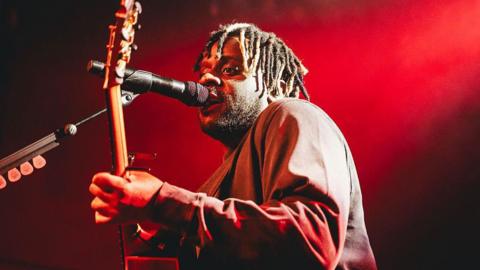 Myles Smith performs during BRITs Week 2026 in Nottingham - red lights surround him on stage as he holds his guitar and sings.
