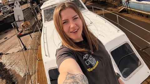 A woman takes a selfie on the bow of a small houseboat. She has long brown hair, tattoos on her arm and wears a grey T-shirt with black leggings and cream croc shoes.