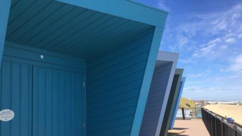 The picture is of a close up of a three modern beach huts in varying shades of blue. The huts take up the half the picture the other half is of the promenade and Lowestoft beach which is sandy.