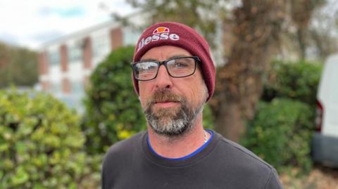 Chris Mollet - A man wearing a burgundy beanie hat, with the brand Ellesse on it, wearing black glasses. He has a brown, black and grey beard.
He's wearing a black jumper, on top a blue shirt and a silver chain. 