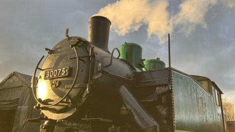 A green and black steam locomotive lit up by the late afternoon sun. The badge on the front bears the number 30075. Steam is coming from the chimney on the top.