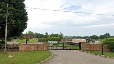 Entrance to the equestrian centre showing wide black metal gates with brick walls on either side. There is a gate and a tree to the left. There are fields behind the gate and some low buildings. There are some clouds in the sky.