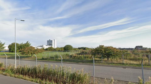 Energy Park Way in Grimsby showing a road with chain link fences running alongside it and fields and factories in the background.