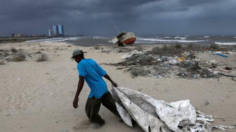 Sooraj, 32, a fisherman and diver, pulls a sheet to cover his belongings, with rain clouds in the background, before the arrival of cyclonic storm