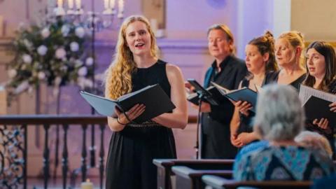 Classical singer Lucy Cox is singing in a choir dressed in black, with other singers stood behind her and to the right.