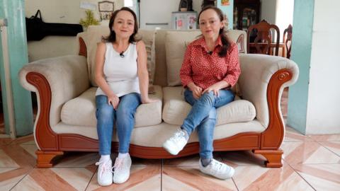 María Luisa sits on a cream sofa with her sister María del Cisne. María Luisa has shoulder-length brown hair and is smiling, wearing a white vest, jeans and trainers. María del Cisne has longer dark hair, with the front pinned back, a red shirt with white polka dots and is also wearing jeans and white trainers.

