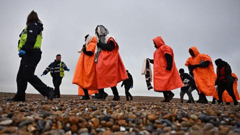 A British Immigration Enforcement officer (L) and an Interforce security officer (2L), escort migrants, picked up at sea by an Royal National Lifeboat Institution (RNLI) lifeboat whilst they were attempting to cross the English Channel, on the shore at Dungeness on the southeast coast of England, on December
