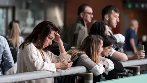 Women stand at a station in Madrid, one looking at their phone and leaning their head on their hand. 