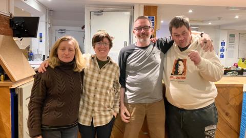 Four people are standing inside the Shrewsbury Ark. Two women and two men have their arms around each other. On the left, the woman has blonde hair and a brown jumper. Next to her, the woman has short brown hair and glasses. One man has short grey hair and glasses, while the other has brown hair and is wearing a cream hoodie.