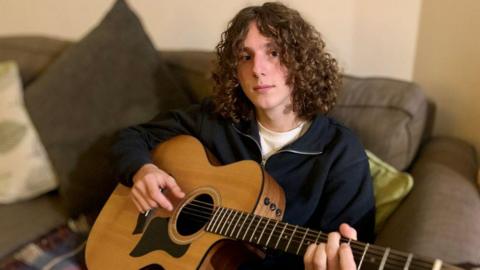 Young man with long curly hair sat holding a guitar on a sofa