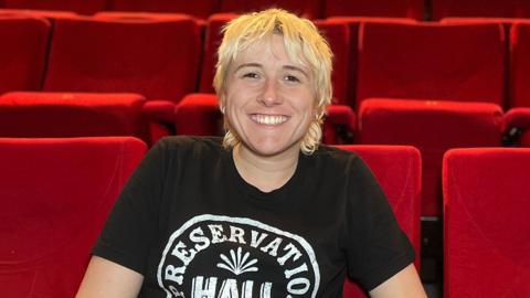 Maisie has short, shaggy blond hair and sits smiling in a theatre auditorium, leaning forward with hands clasped. She is wearing a black T‑shirt and sits among rows of bright red seats that fill the background.