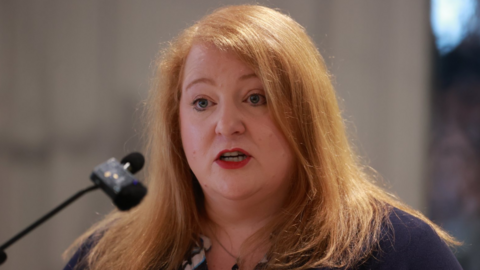 Naomi Long is standing in front of a microphone, She has ginger hair and is wearing red lipstick.