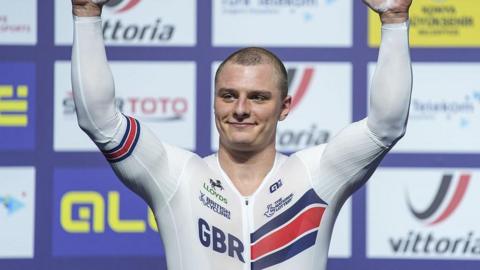 Matt Richardson in white British kit with GBR lettering raises arms aloft in front of sponsor background