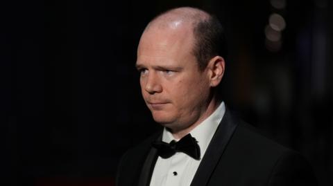 Gordon Lyons is wearing a black and white tuxedo suit. His bow tie and jacket are black. His shirt is white. The background is dark and blurred. He's looking away from the camera. 