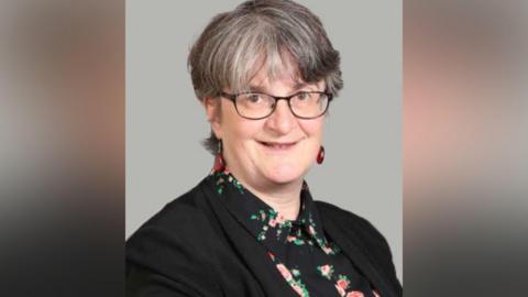 A woman with short black and grey hair looks into the camera, wearing black-rimmed glasses. She has a dark jacket on over a black shirt with a light floral pattern.