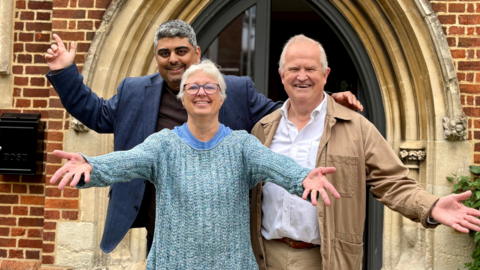 Three people - two men and a woman - stood outside a church with their hands in the air in a celebratory way