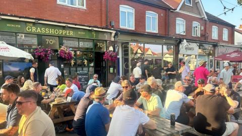 Crowds sit outside in the sun on chairs table and chairs set outside The Grasshopper pub.