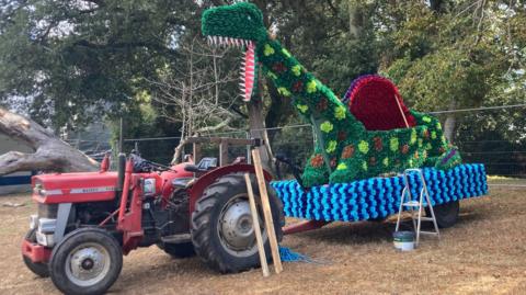 A colourful float appears to be under construction in a park or showground, with a large green dinosaur-like figure covered in paper flowers mounted on a trailer. 