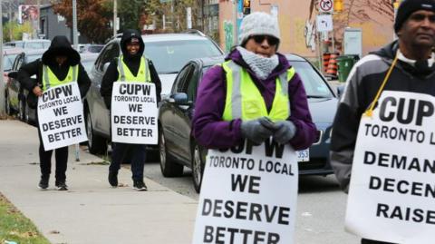 An image of striking Canada Post workers in Toronto, holding signs that say "TORONTO LOCAL WE DESERVE BETTER" as they walk on the sidewalk next to a busy Toronto street