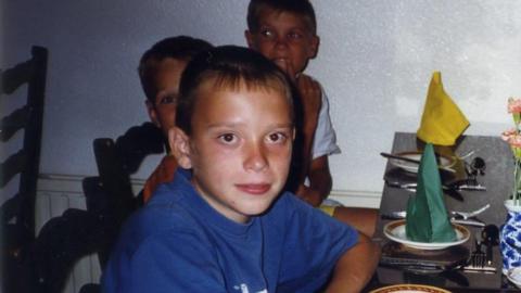 Anton wearing a blue t shirt on the left. There are several other people smiling at the camera as they sit down at a dinner table. There are green and yellow napkins on plates.
