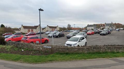 Rock Street Car Park in Thornbury. It is situated on a hill, on a bend in the road. It is surrounded by a brick wall and a pavement.