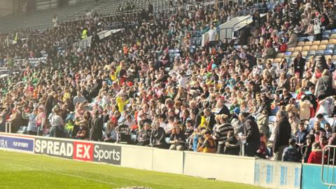 A crowd of people in the stands of a football pitch. Mostly young children with their guardians, many of them are in fancy dress of characters from children's books.