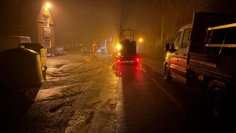 A street lit by streetlamps is flooded. It is dark but the lights of several maintenance trucks can be seen.