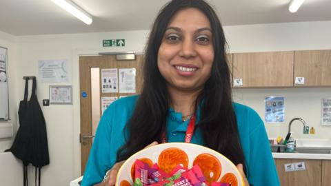 A woman wearing a blue top, smiling and looking directly at the camera. She is stood in the classroom. 