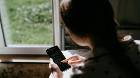 Stock image of a young woman using a phone. Her back is to the camera and we are looking over her left shoulder at her phone screen. A window is in the background.