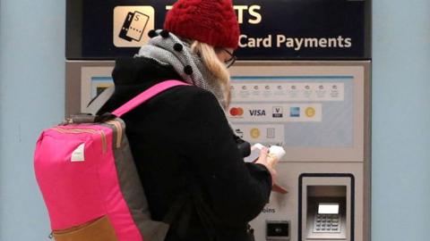 A woman wearing a red hat, a grey scarf and a black jacket with a bright pink rucksack buys a train ticket at a self-service machine in Waterloo station, London