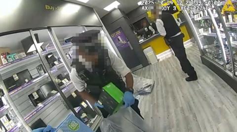 Two police officers in an electronics shop. One pictured placing a green box into a plastic sack. He is wearing a short-sleeved white shirt, a police gilet and blue gloves. Both officers' faces are pixelated. The shop floor and walls are grey.