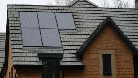 A close up view of a tiled roof on a house. It is a red brick house with black tiles on its roof. It also has five solar panels installed on the roof. Two windows on the front of the house can also be seen.