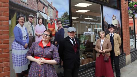 Four members of staff from Beamish Museum wearing old-fashioned clothing stand in front of the Beamish Emporium