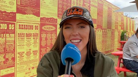 Artist LYVIA has long brown hair and wears a baseball cap and a green jacket. She's holding a newsbeat mic