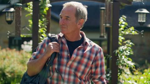 Robson Green walking through a green space with a rucksack over his shoulder