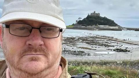 Matt Saunders is standing in from of St Michaels' Mount in Penzance. He is wearing glasses and a white cap. He has a light brown beard and moustache.