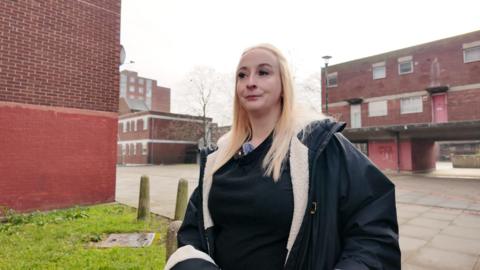 Danielle, a blonde woman in a winter coat, looks wistfully away from the camera against the backdrop of a red-brick housing estate