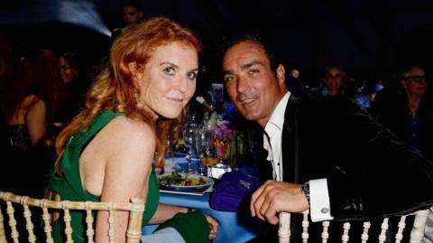 Sarah Ferguson and Manuel Fernandez sit at a table laden with glasses, plates and a blue table cloth. She wears a green dress, he wears a black velvet jacket and a white shirt open at the neck. They sit at wickerwork-style chairs, and are looking over their shoulders at the camera