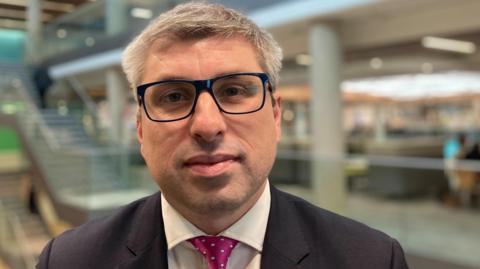 Mark Arnull, who has short hair, black-framed glasses and a dark jacket over a white shirt and pink tie. He is standing in an atrium in an office building.