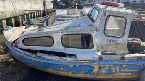 A derelict looking small boat in a harbour while the tide is out. It is white and blue with patches of badly flaking yellow paint on the hull. and a life ring on its roof.