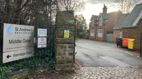 Entrance and gate lodge at a mental health hospital. In the foreground to the left are a number of signs. A cobbled road can be seen as well as multi-coloured bins. There are trees and a grey sky.
